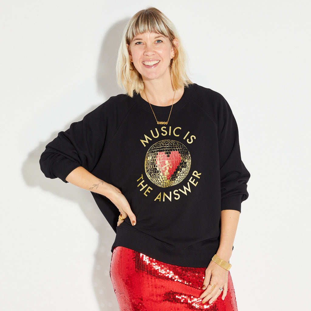 Woman wearing a black sweatshirt with 'Music is the Answer' text and a disco ball graphic, standing against a white background.