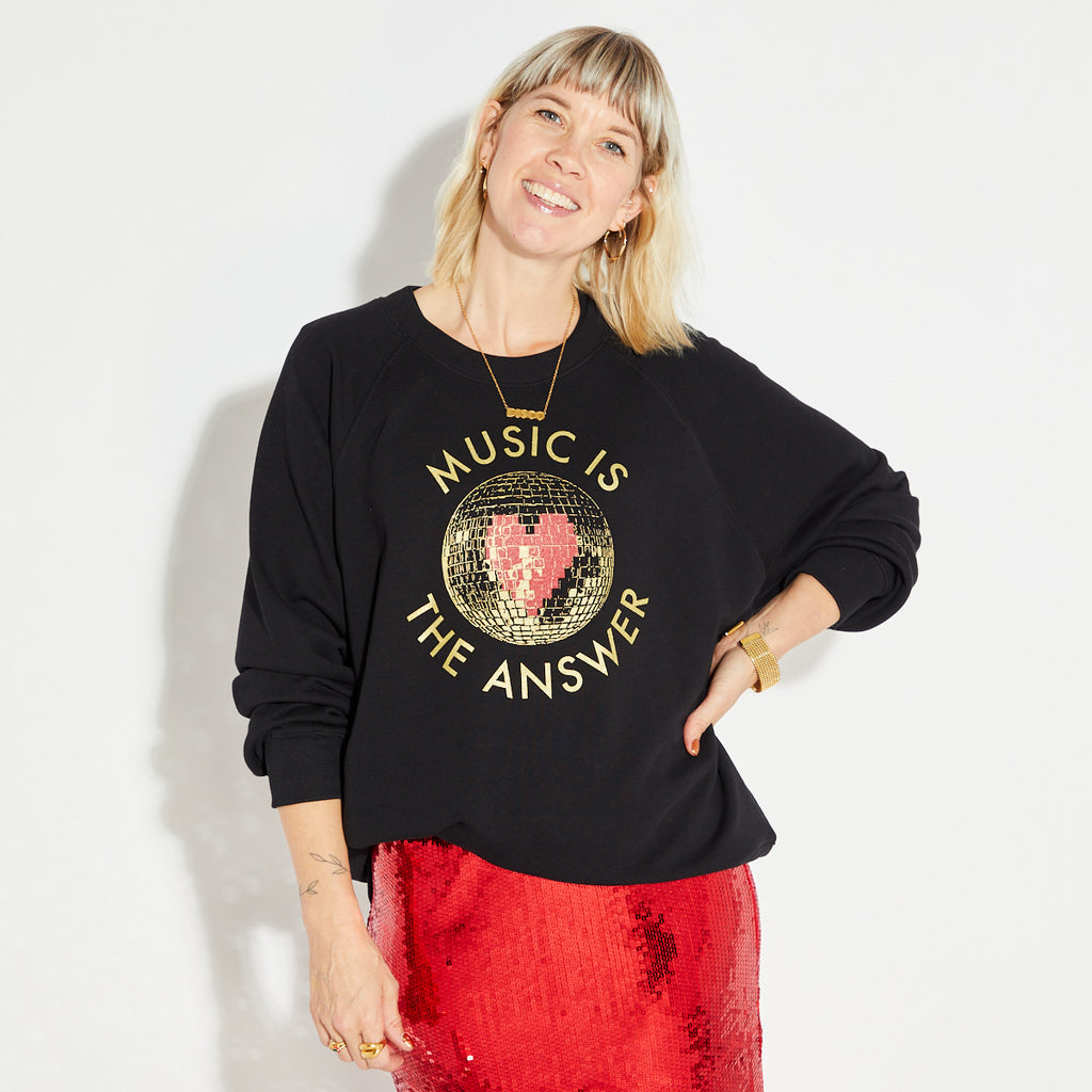 Woman wearing a black sweatshirt with 'Music is the Answer' text and a disco ball graphic, standing against a white background.