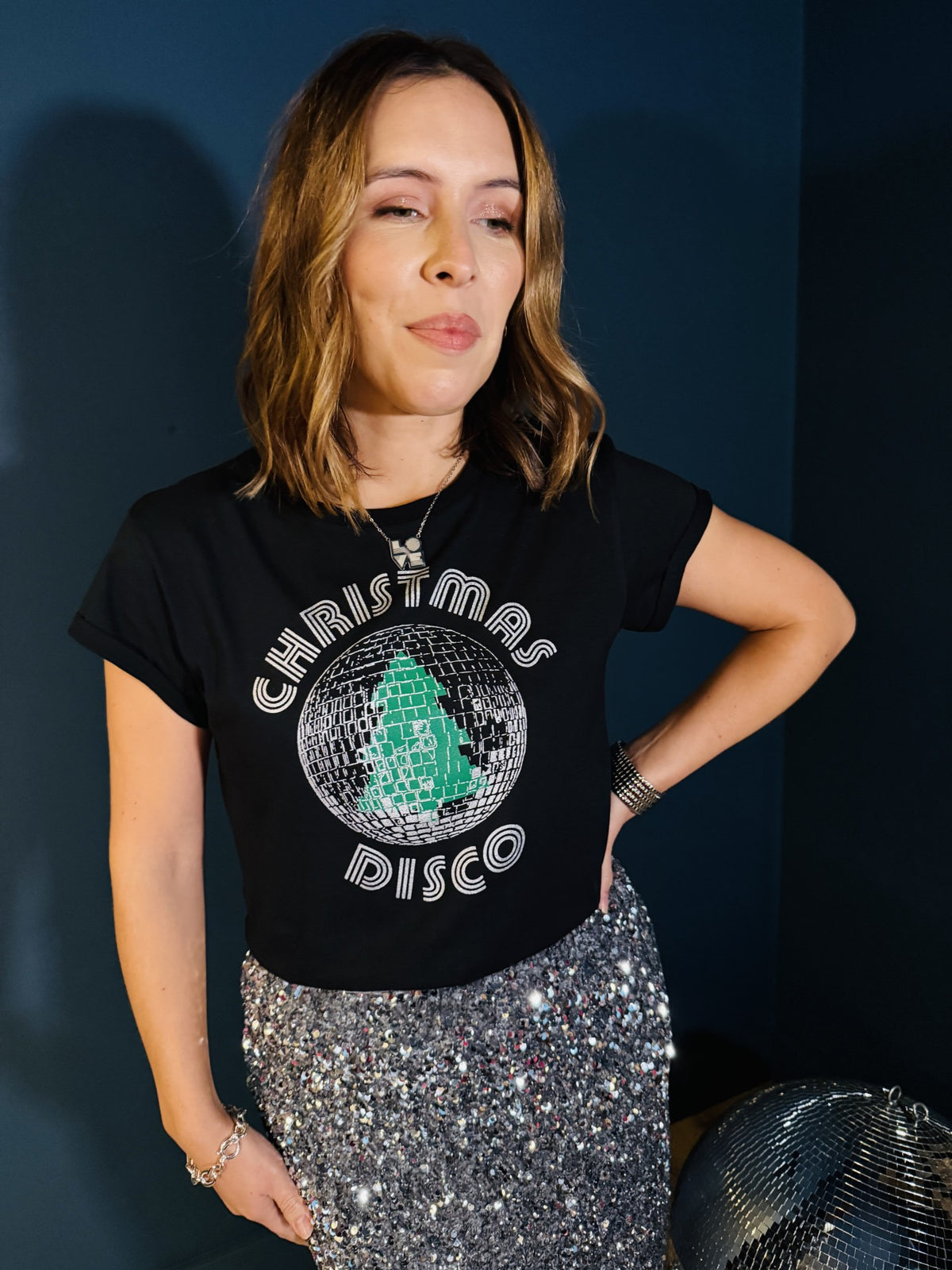 A woman wearing a black graphic t-shirt with a silver and green 'Christmas Disco' disco ball design, paired with a silver sequined skirt.
