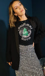 A woman wearing a black t-shirt with a green Christmas disco graphic, styled with a black blazer and a silver sequined skirt, posing against a dark blue wall.