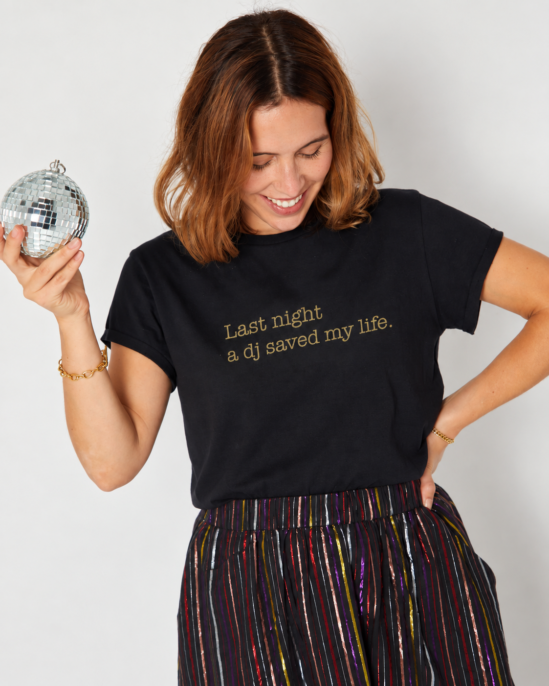 A woman wearing a black graphic t-shirt with gold text that reads 'Last night a dj saved my life.' She is holding a silver disco ball and wearing a colorful striped skirt against a plain light gray background.