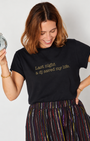 A woman wearing a black graphic t-shirt with gold text that reads 'Last night a dj saved my life.' She is holding a silver disco ball and wearing a colorful striped skirt against a plain light gray background.