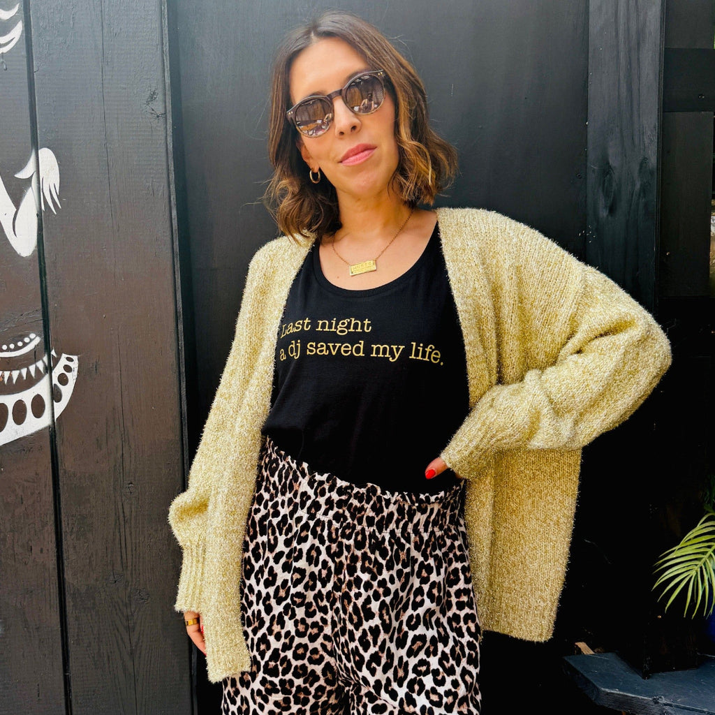 Woman wearing a beige cardigan, black t-shirt with text, and leopard print pants against a dark background.