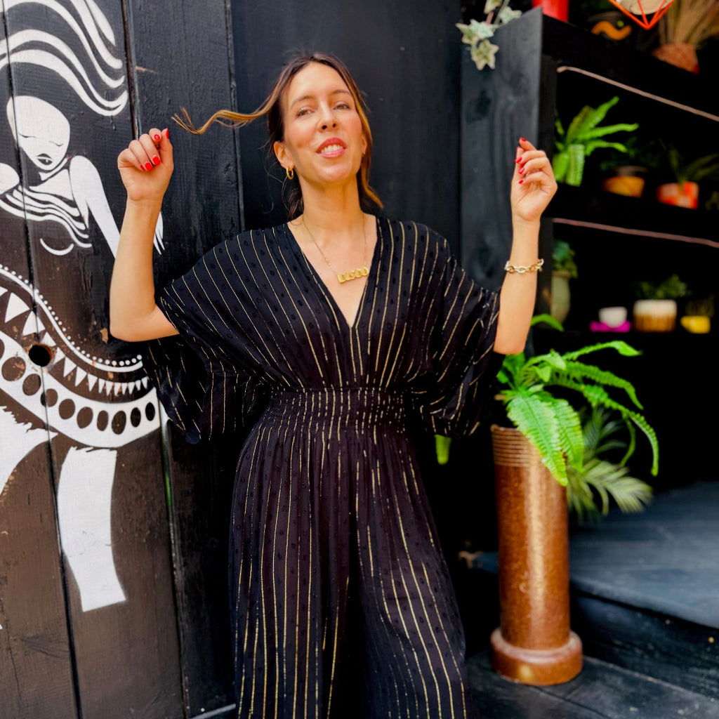 Woman in a black pinstripe jumpsuit posing in front of a decorative wall with plants.