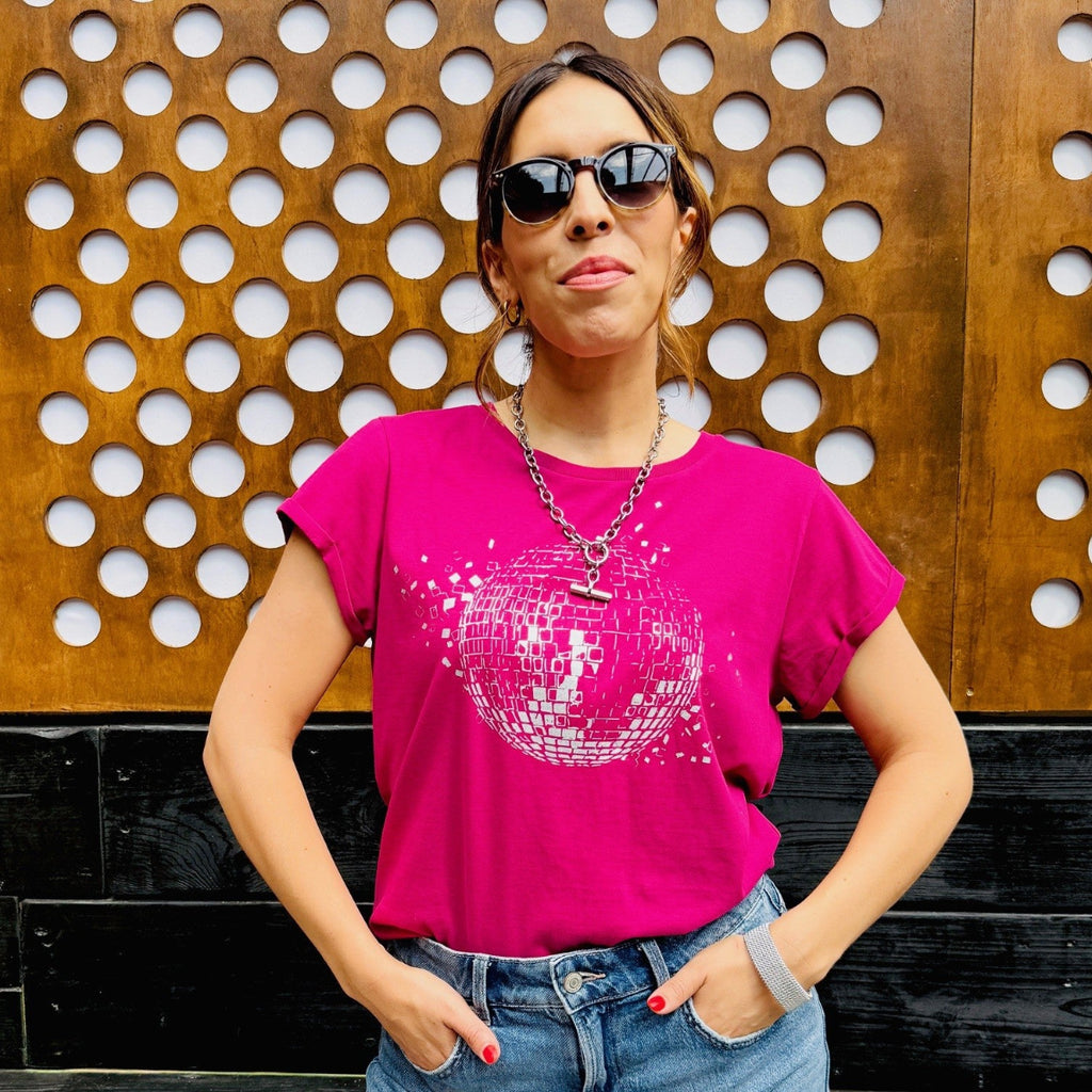 Woman wearing a pink t-shirt with a disco ball design, sunglasses, and denim shorts against a wooden wall.