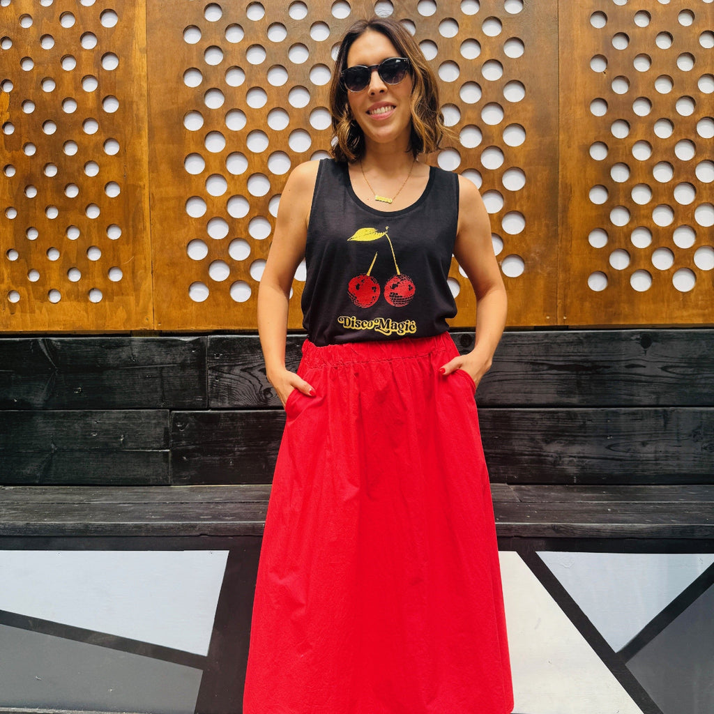 Woman wearing a black tank top with red disco magic cherries and a red skirt standing in front of a wooden wall.