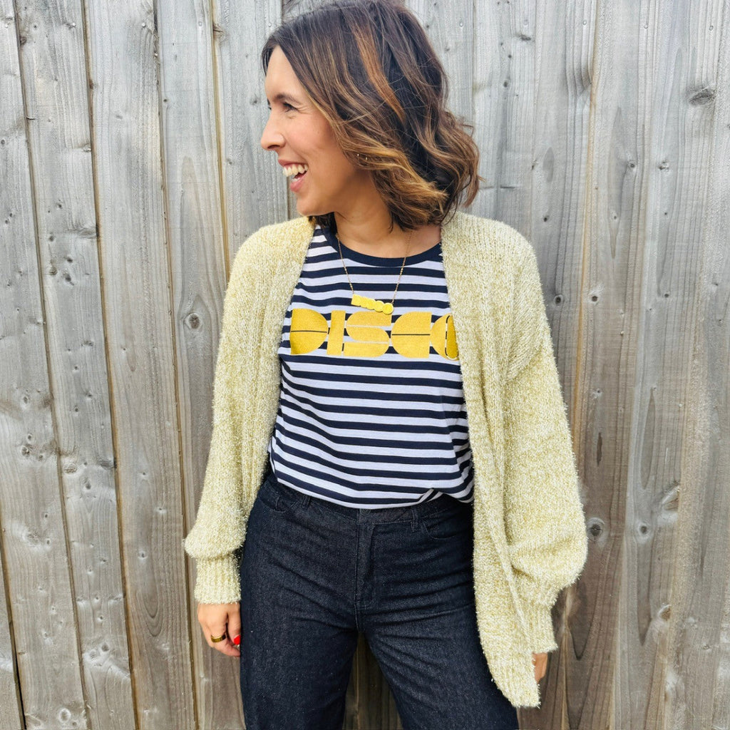 Woman wearing a striped shirt with gold 'DISCO' text and a light cardigan against a wooden fence.