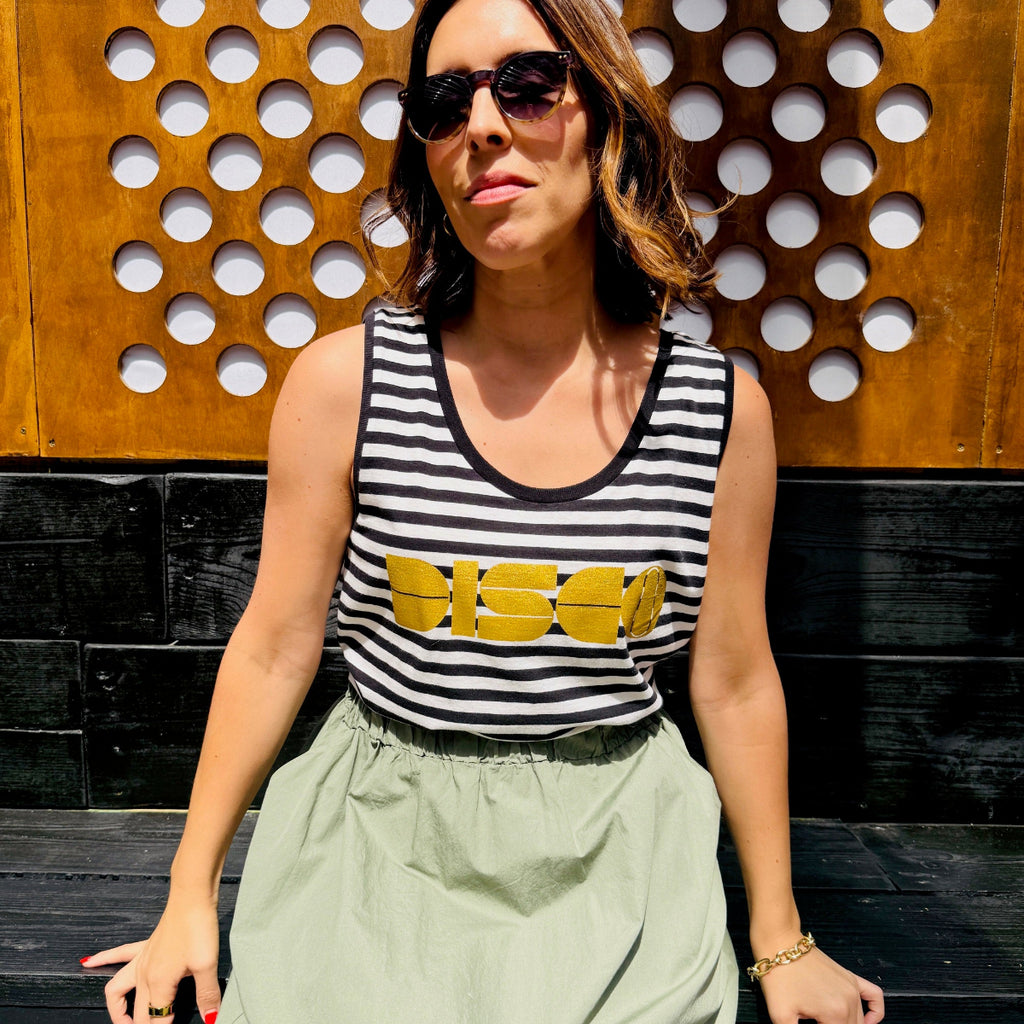 Woman wearing a black and white striped tank top with yellow text, sitting against a wooden and black stone wall.