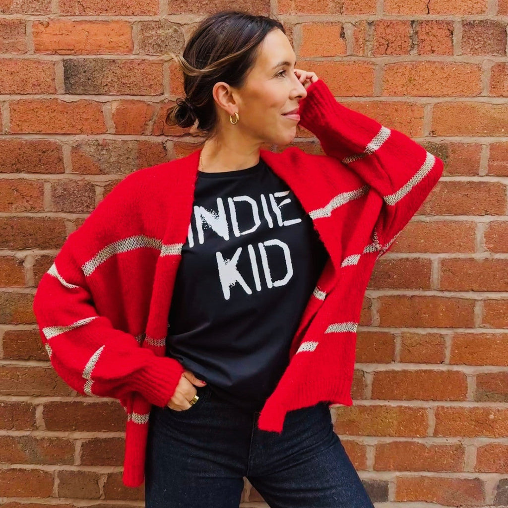 Woman wearing a red cardigan over a black t-shirt with 'INDIE KID' text, standing against a brick wall.