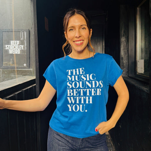 Woman wearing a blue t-shirt with 'music sounds better with you' text, standing indoors.