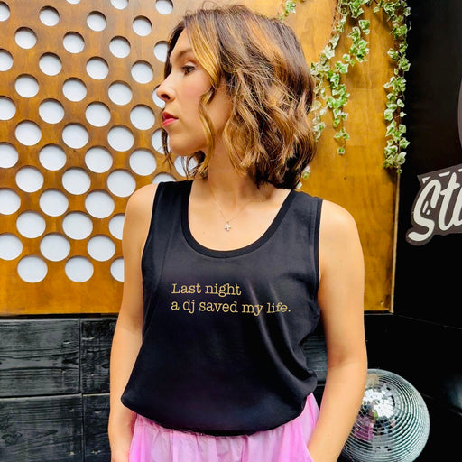 Woman wearing a black tank top with last night a dj text, standing in front of decorative wall and disco ball.