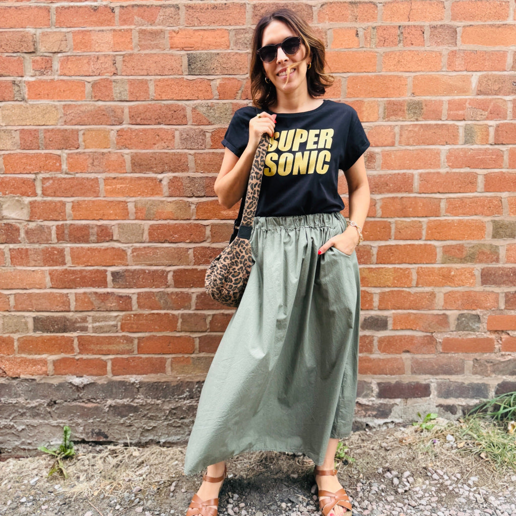 Woman wearing a black t-shirt with 'Supersonic' text, green skirt, and sunglasses against a brick wall.
