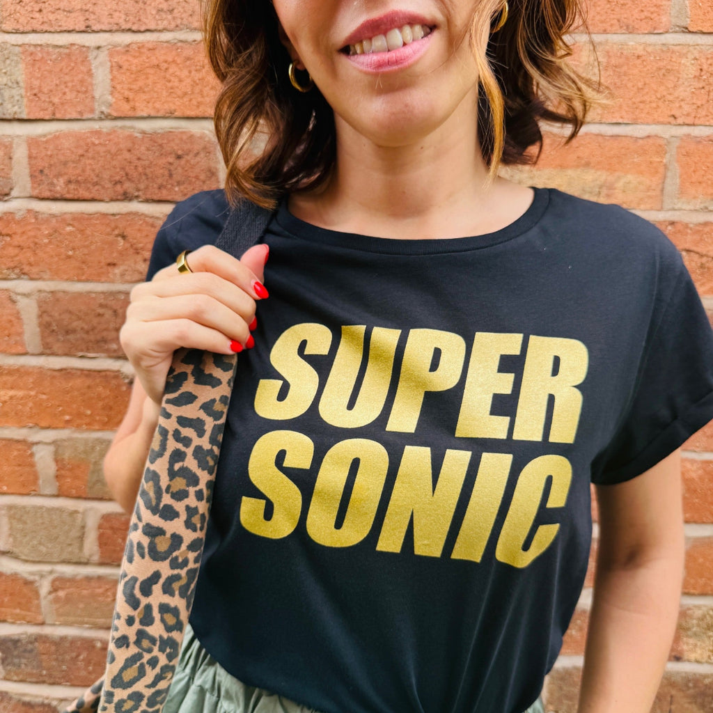 Person wearing a black t-shirt with 'SUPERSONIC' in gold letters against a brick wall.