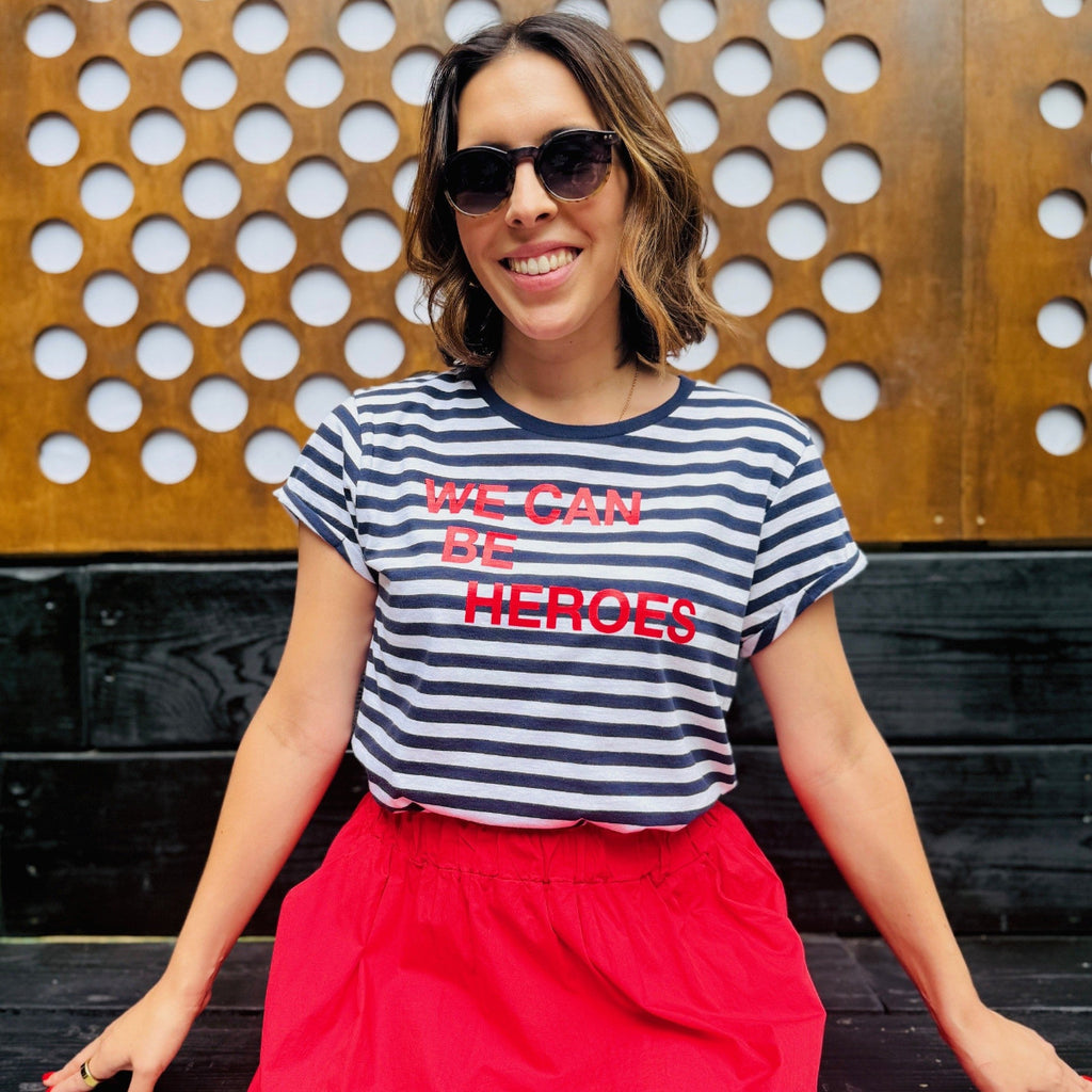 Woman wearing a striped shirt with 'WE CAN BE HEROES' text, sitting against a wooden wall.