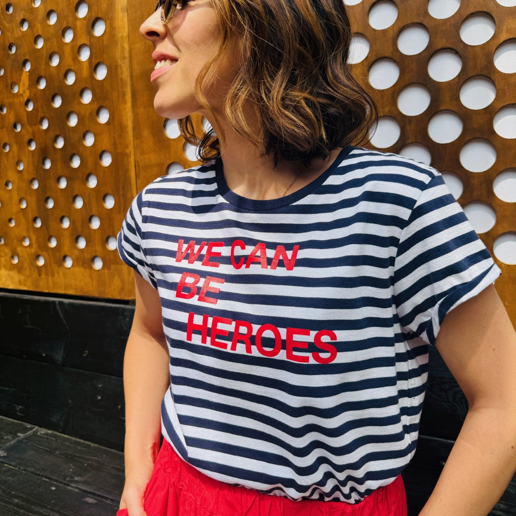 Person wearing a striped shirt with 'WE CAN BE HEROES' text against a perforated metal wall.